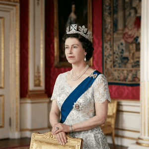 Queen Elizabeth II wearing a jeweled crown, pearl necklace, blue sash, and white gown in an ornate room